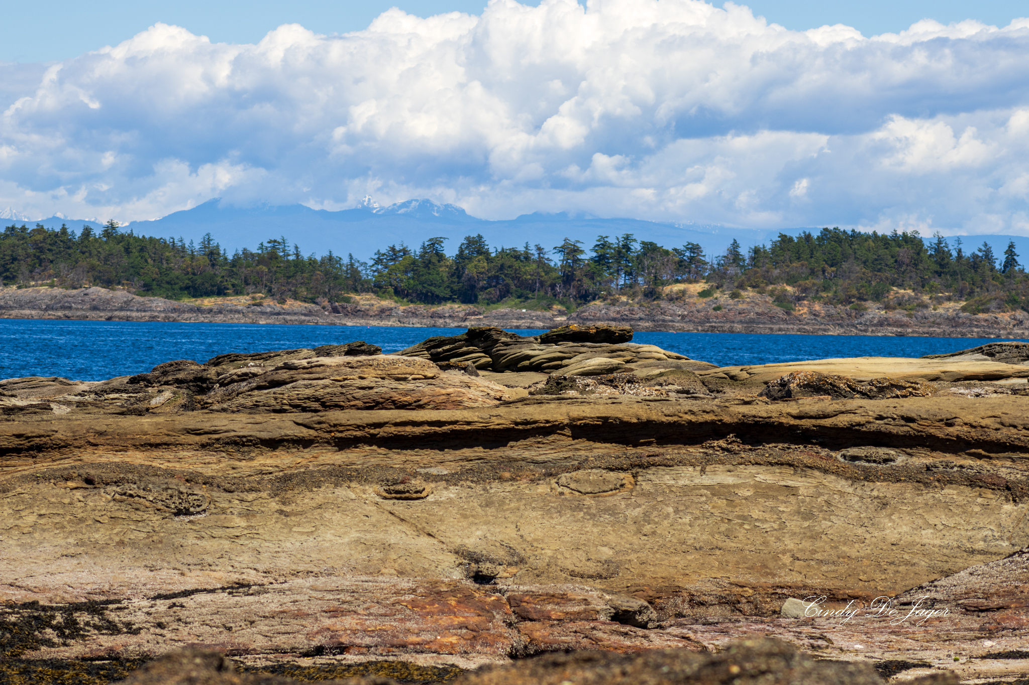Madrona Park view of layered shoreline rocks, calm blue ocean, a forested island, and distant mountain peaks.