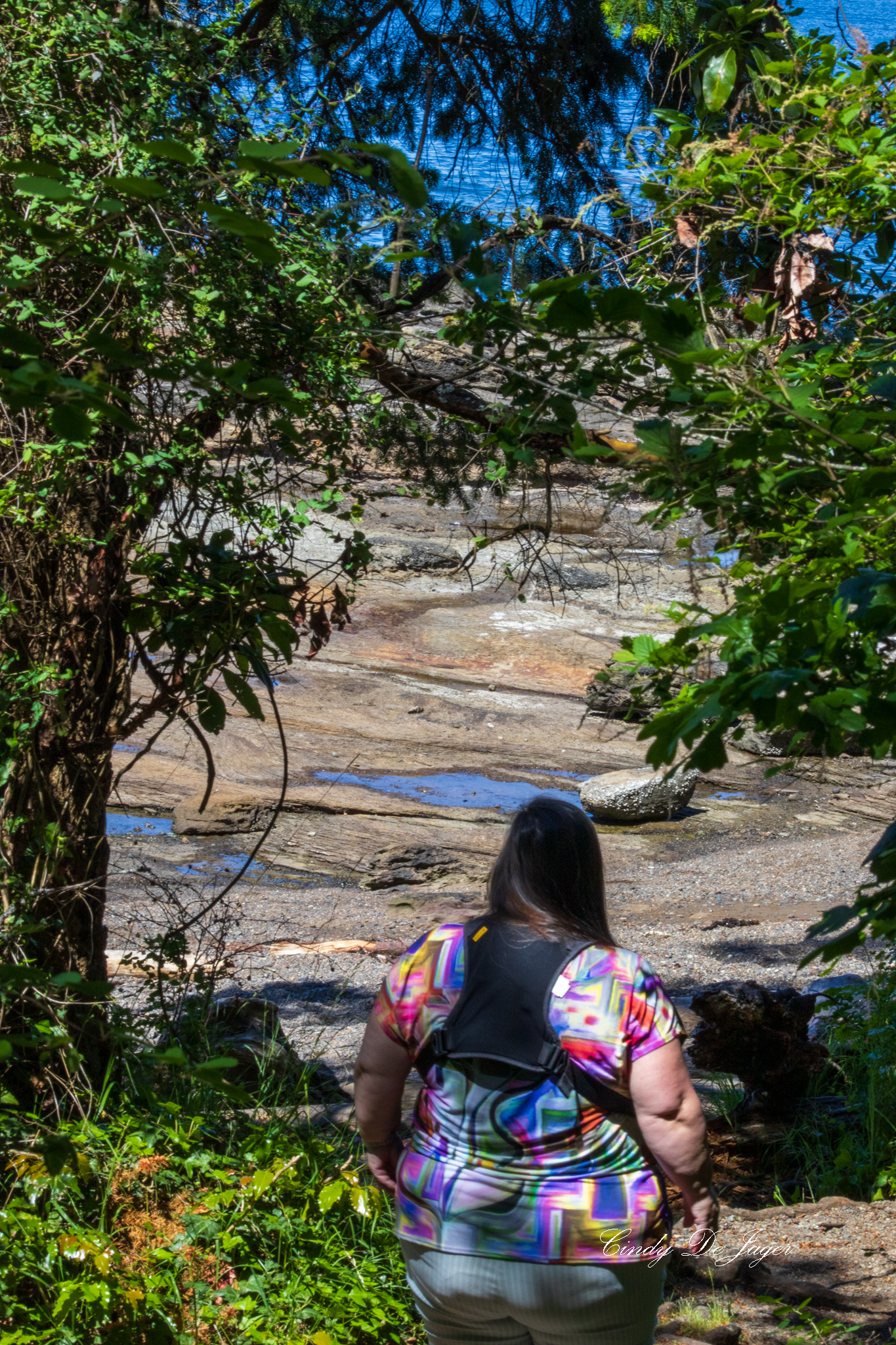 Person standing on a rocky shoreline at Madrona Park, surrounded by dense green foliage and looking out toward the blue water.